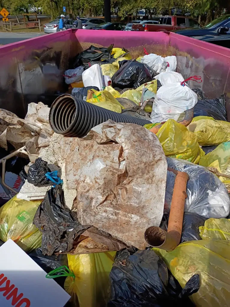 Pink dumpster heaped with bags of trash, a black drainage pipe, a rusty pipe and muddy plastic sheeting pulled from the river.