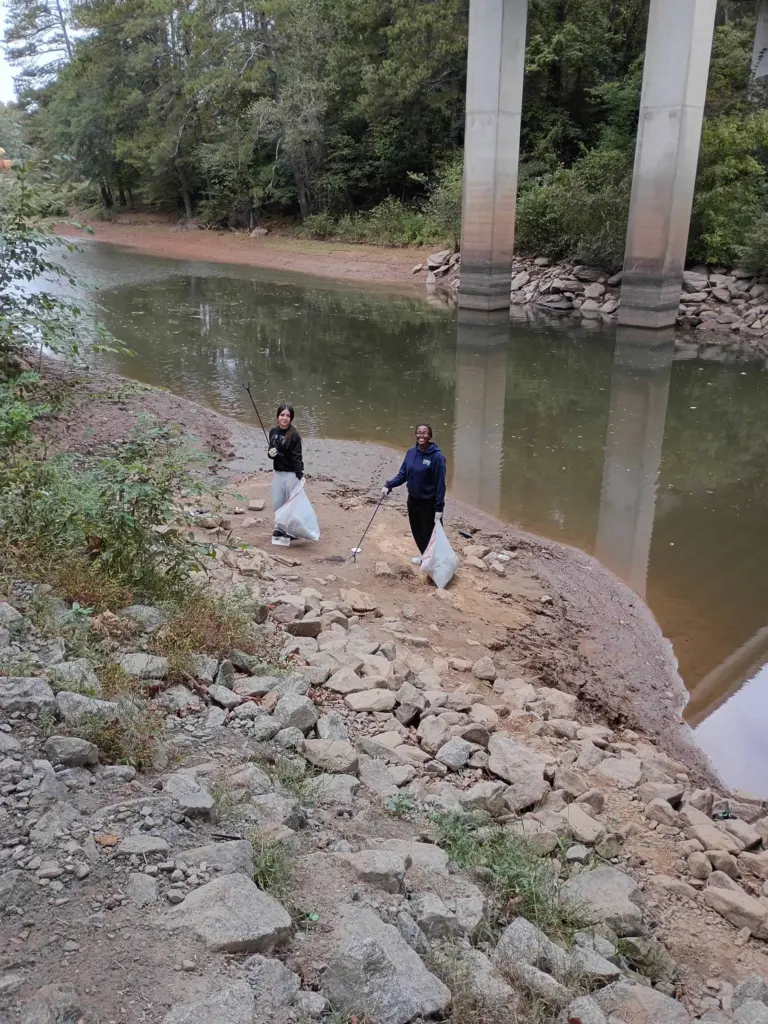 Two volunteers with trash grabbers and full bags stand on a sandy riverbank beneath the concrete pillars of a highway bridge.