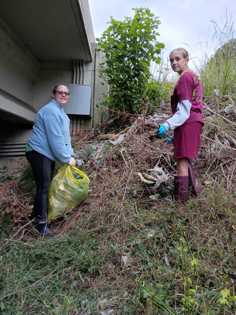 Two volunteers in gloves pick litter out of tangled brush and tall grass on a slope beside a concrete bridge.