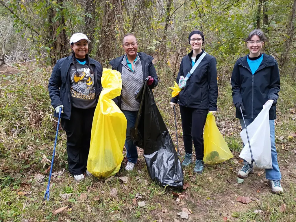 Four smiling volunteers in jackets hold trash grabbers and yellow, black and white bags on a wooded trail.