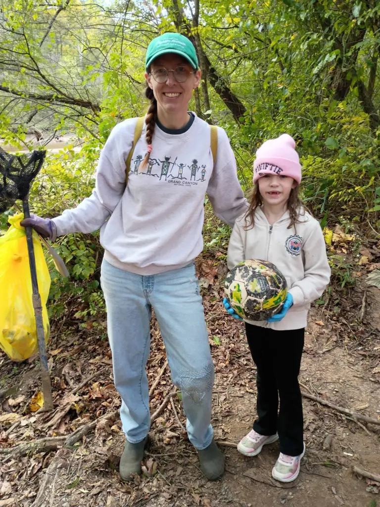 Smiling adult volunteer with a net and yellow trash bag stands beside a child holding a muddy soccer ball found by the river.