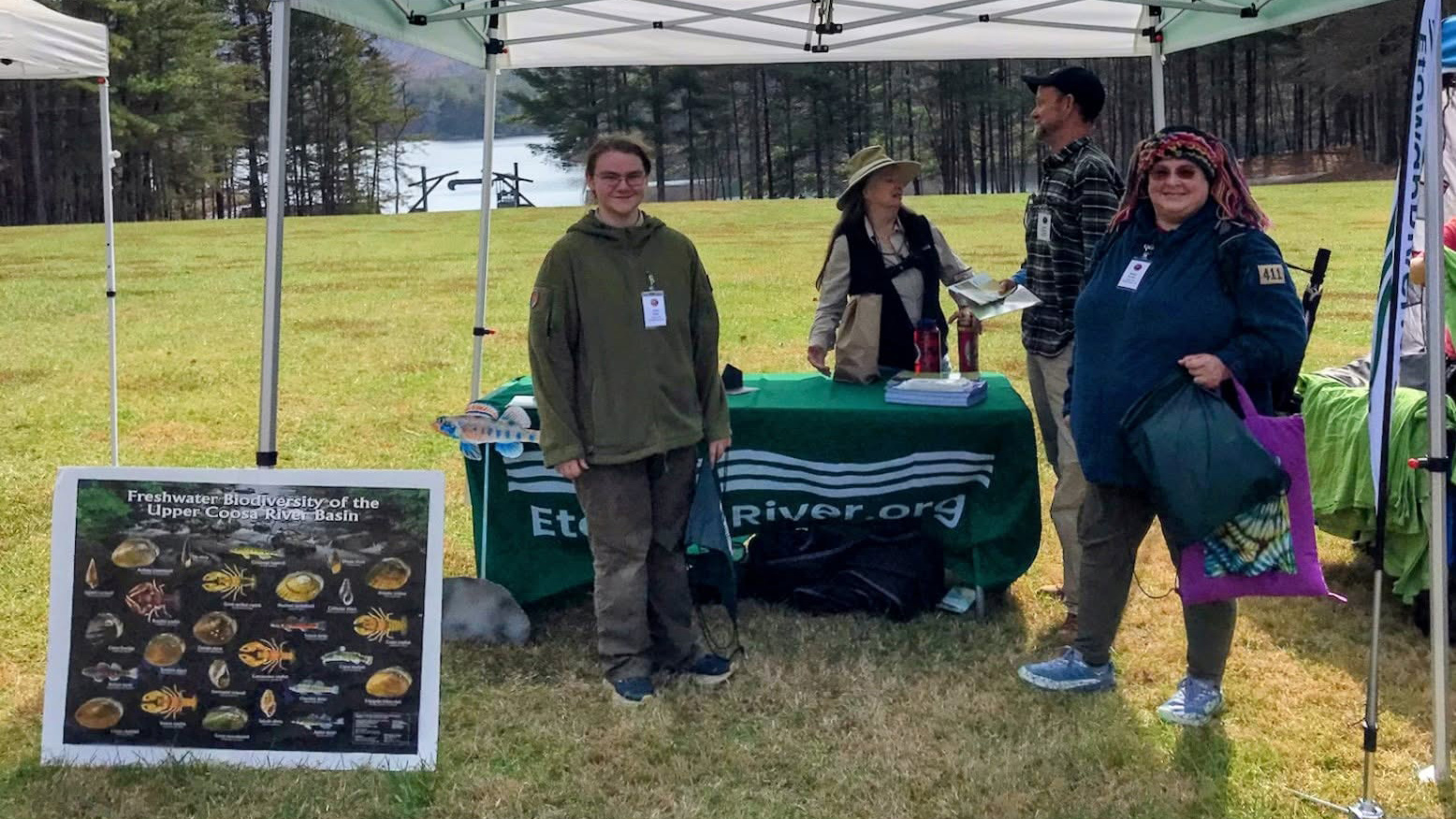 Upper Etowah River Alliance volunteers at an outdoor booth with a green EtowahRiver.org table, beside a "Freshwater Biodiversity of the Upper Coosa River Basin" poster, on a grassy field with a lake and pine trees behind them.