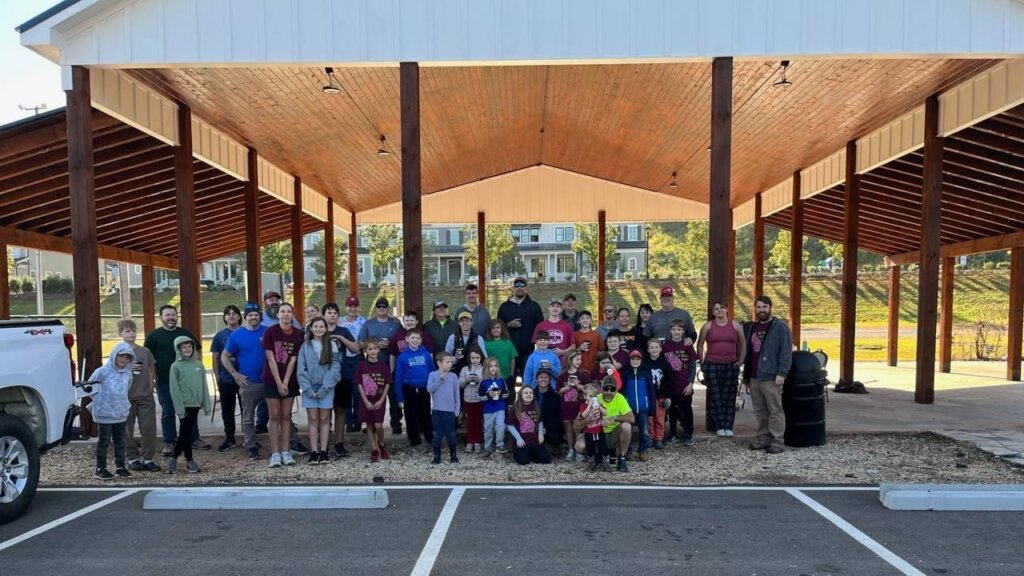 A large group of adults and kids, many in matching maroon T-shirts, poses under a wooden park pavilion before a cleanup.