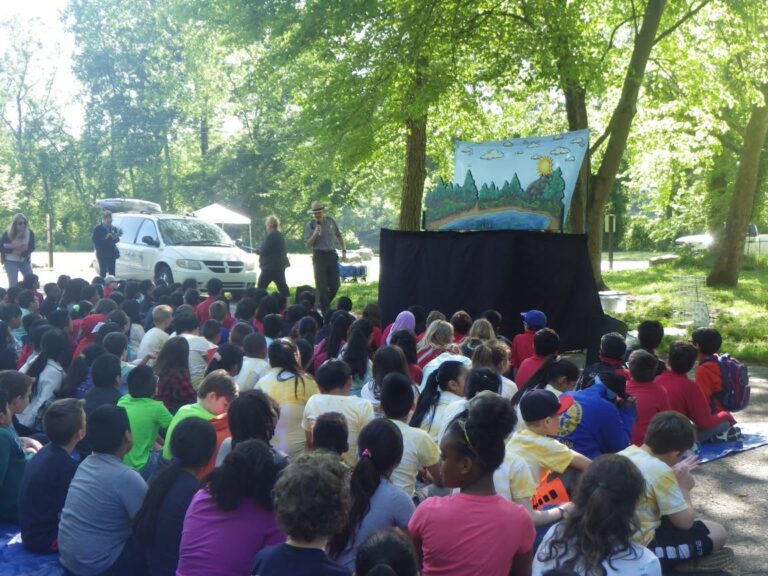 ▎ A park ranger in uniform speaks into a microphone beside a black puppet stage with a painted river-and-forest backdrop, while a large group of children sit on the ground outdoors in a shady, wooded area, watching.