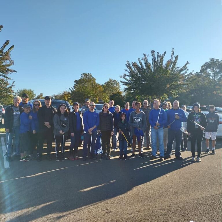 ▎ A group of about 25 volunteers, adults and children, many in matching blue shirts, pose together in a parking lot on a sunny morning. Several hold litter grabbers for a cleanup event.