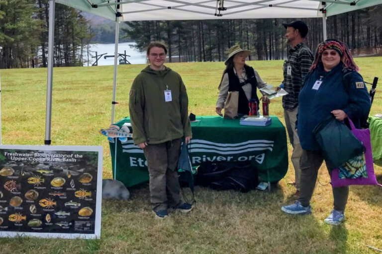 Upper Etowah River Alliance volunteers at an outdoor booth with a green EtowahRiver.org table, beside a "Freshwater Biodiversity of the Upper Coosa River Basin" poster, on a grassy field with a lake and pine trees behind them.