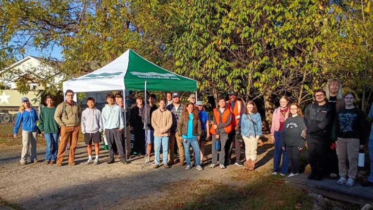 About 25 volunteers of all ages pose in morning sun beside a green and white EtowahRiver.org canopy tent before the cleanup.