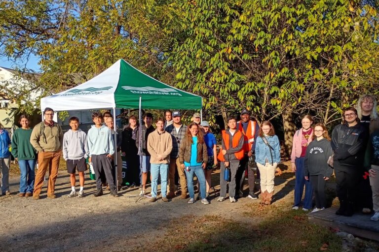 About 25 volunteers of all ages pose in morning sun beside a green and white EtowahRiver.org canopy tent before the cleanup.