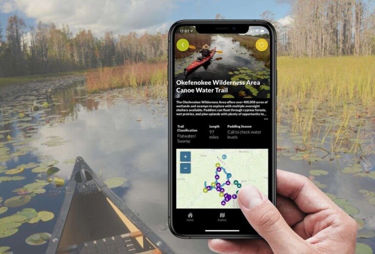 A hand holds a phone showing the Okefenokee Wilderness Canoe Trail in the Georgia River Guide app, in front of a canoe on a lily-pad swamp